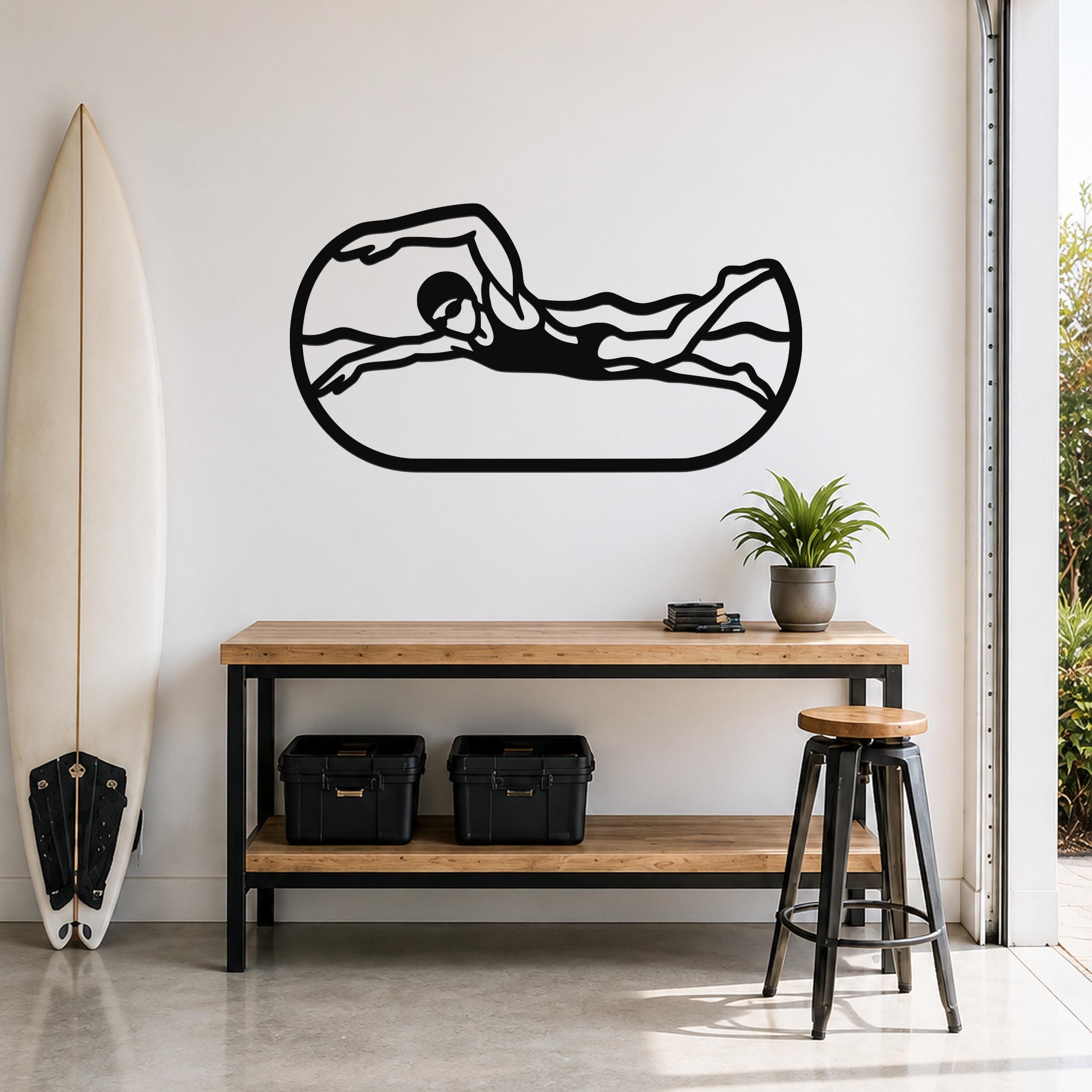 Black metal swimmer wall art with motion-line cutouts, mounted above a wooden console table in a bright room with a surfboard and plant.