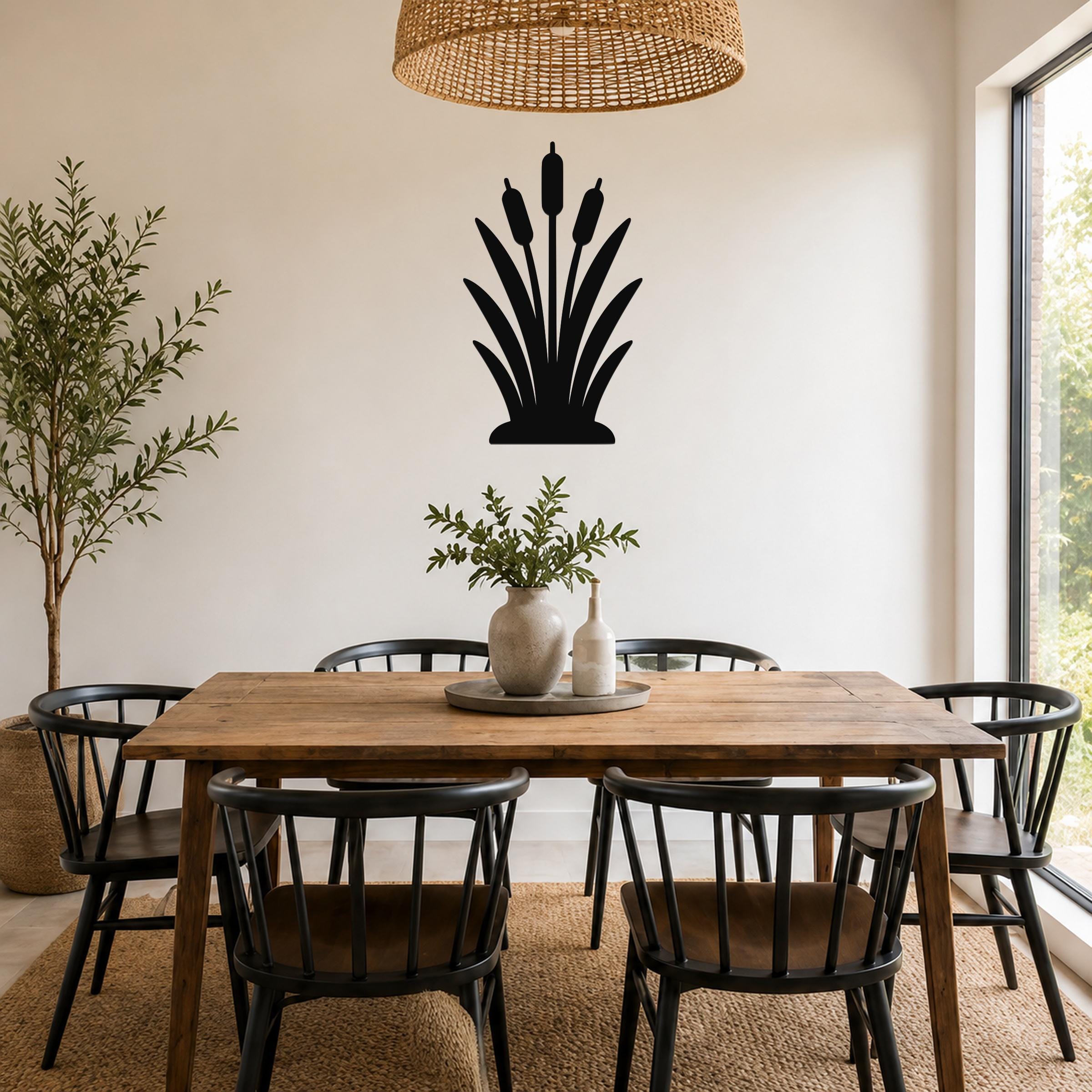 Black metal cattail wall art silhouette with three seed heads and leafy base, mounted on a white wall above a wooden dining table in a bright dining room.