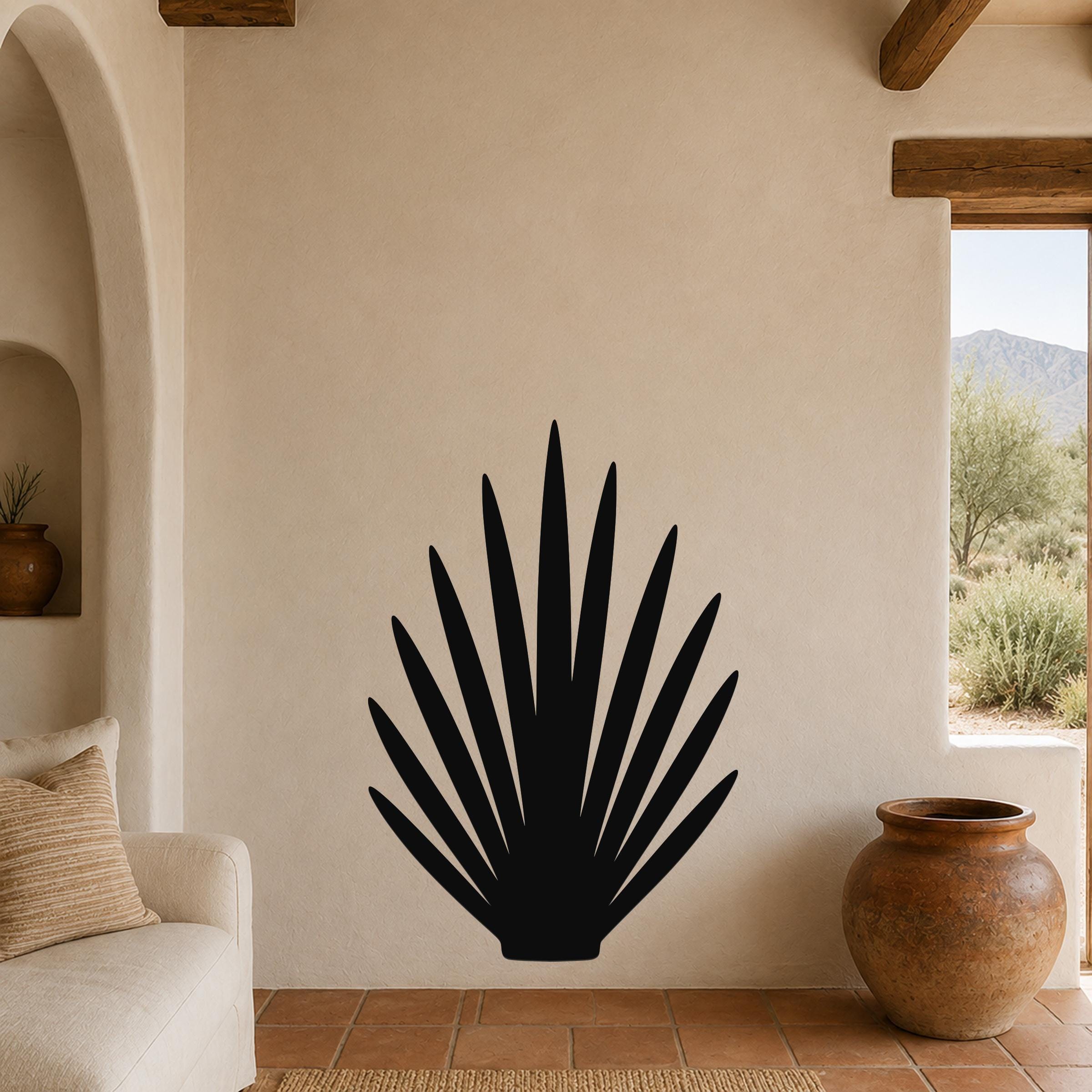 Black metal wall art in a fan-shaped agave silhouette, mounted on a beige stucco wall in a living room with terracotta flooring and wooden beams.