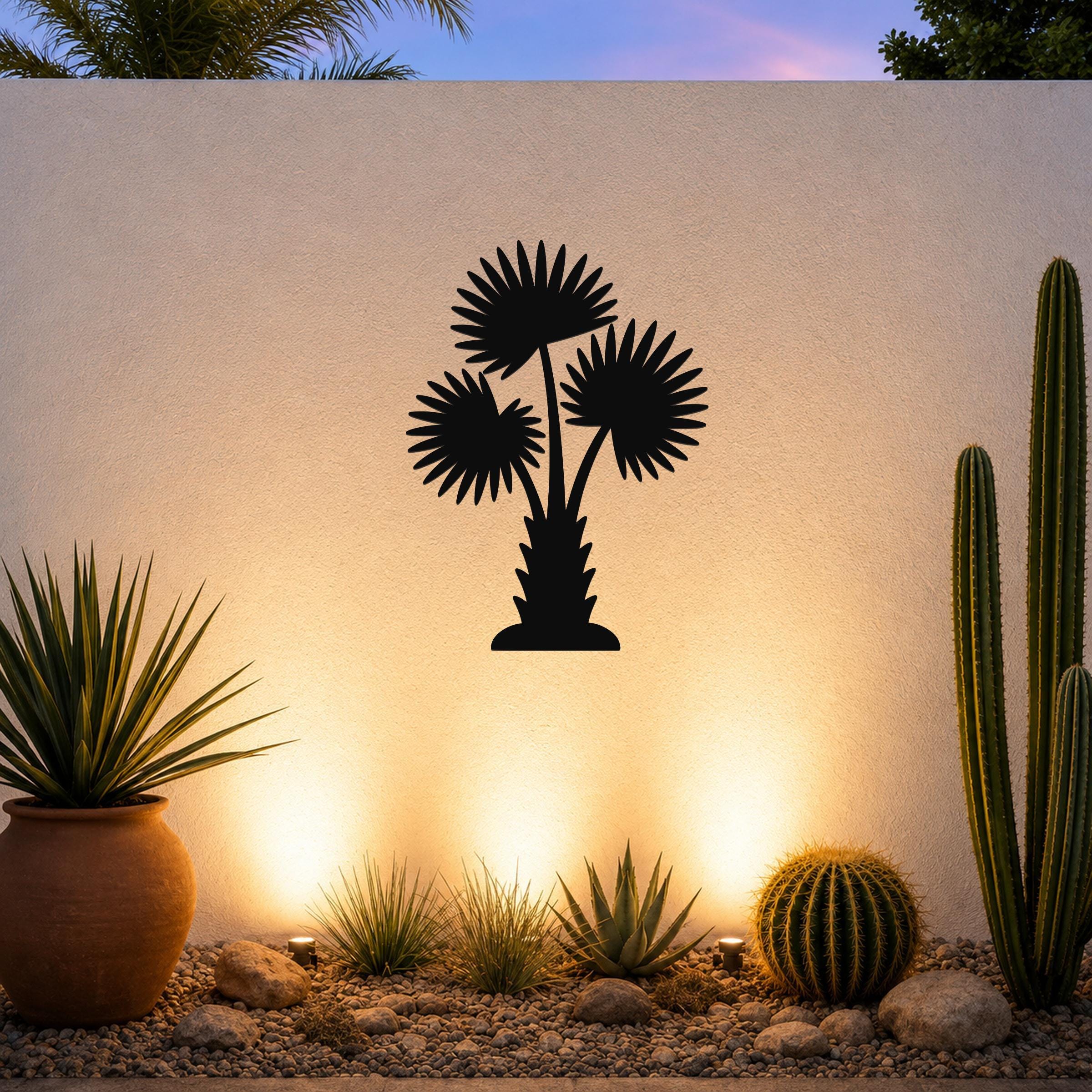 Black metal palm tree wall art with fan-shaped fronds and a textured trunk, mounted on an outdoor wall above a desert garden with cacti and warm uplighting.