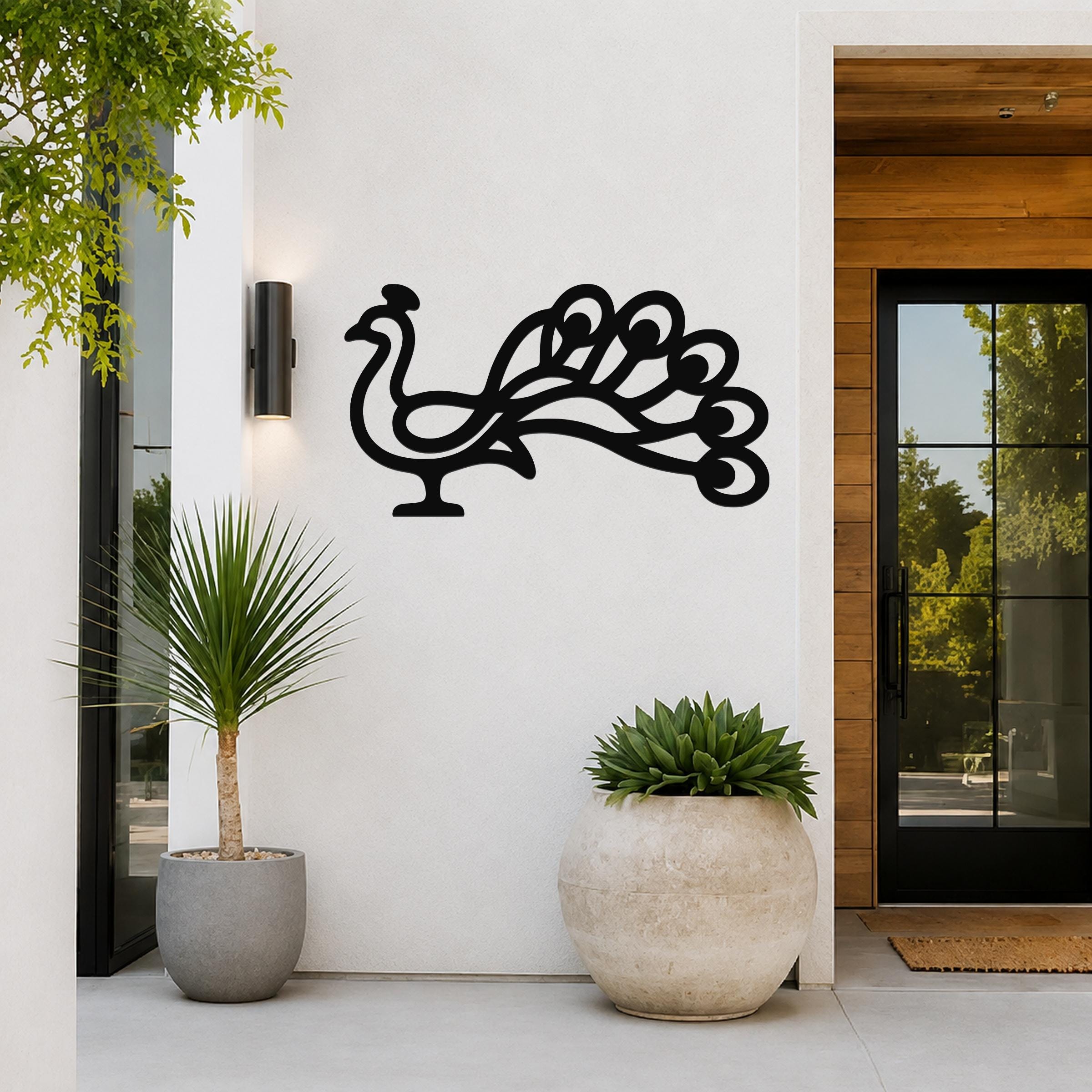 Black metal peacock wall art with a cutout silhouette of a fanned tail, mounted on a white exterior wall beside a glass door and potted plants.