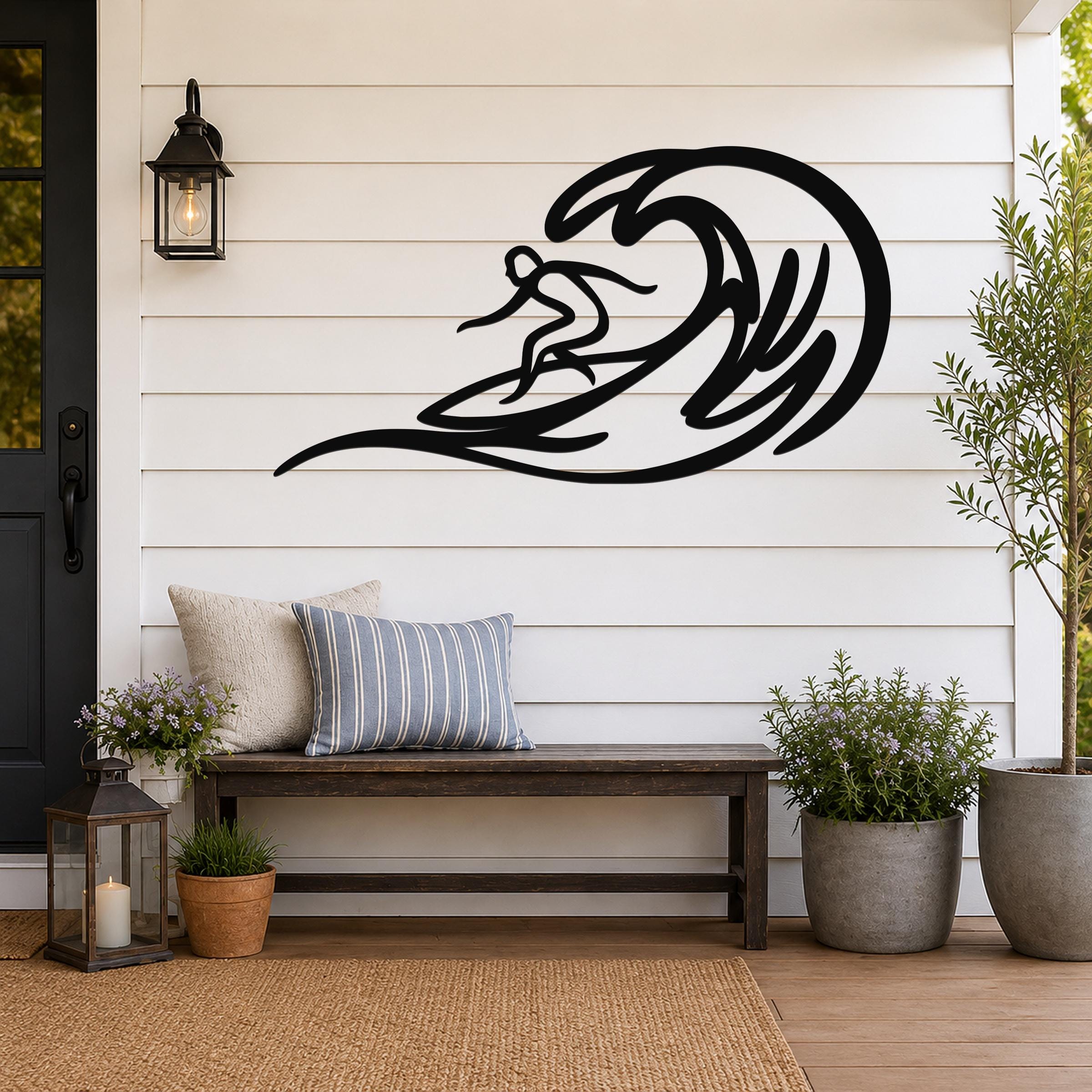 Black metal surfer wall art with wave cutout, mounted on a white shiplap wall above a wooden bench on a porch.