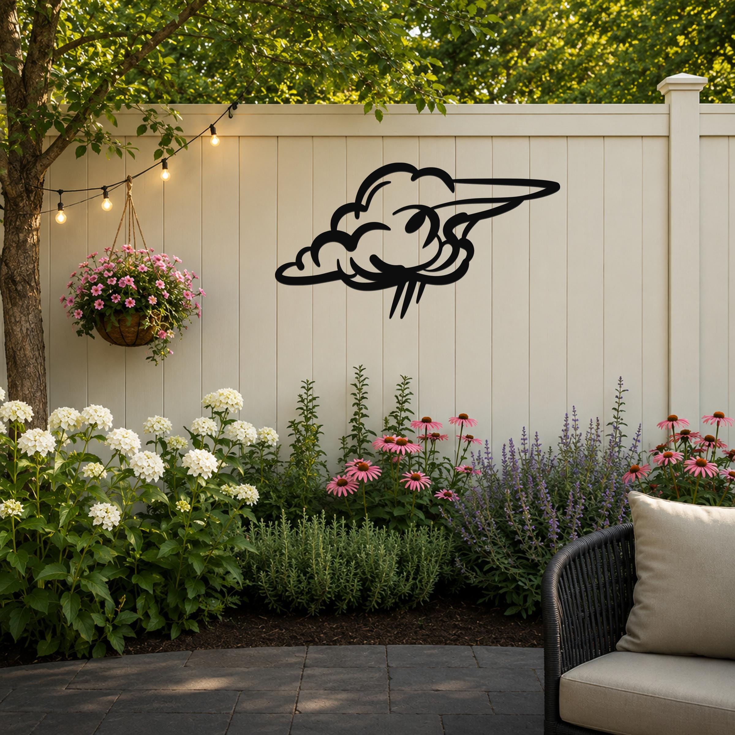 Black metal cloud wall art with a wavy, flowing silhouette, mounted on a light beige outdoor fence above a garden bed with flowers and a hanging planter.