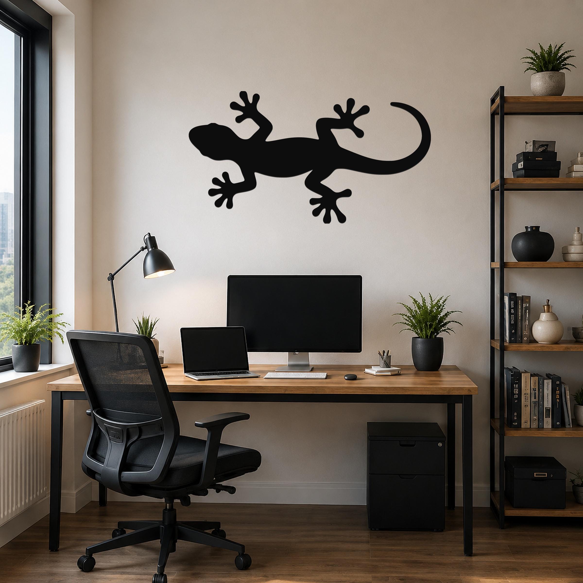 Black metal lizard wall art silhouette mounted above a wooden desk in a home office, featuring a cutout design of a gecko with spread limbs and a long tail.